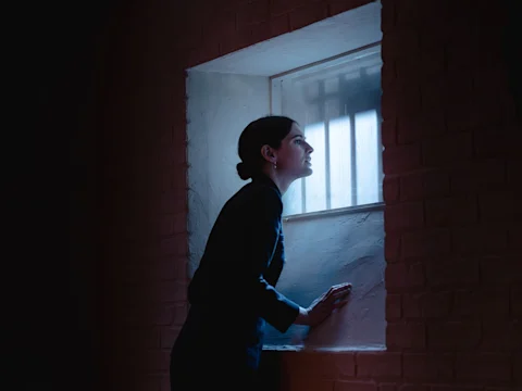 A person with tied-back hair looks out a barred window in a dimly lit room, with their hands resting on the windowsill.