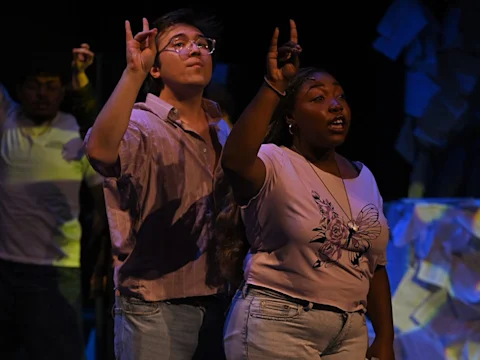 Two people on stage, mid-performance, hold one hand up with pinky and index fingers raised. They appear to be singing or speaking. Background is dark with some paper-like set pieces visible.