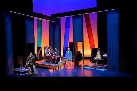 Four musicians perform on stage with saxophone, drums, bass, and piano, surrounded by colorful, geometric set pieces and dramatic lighting.