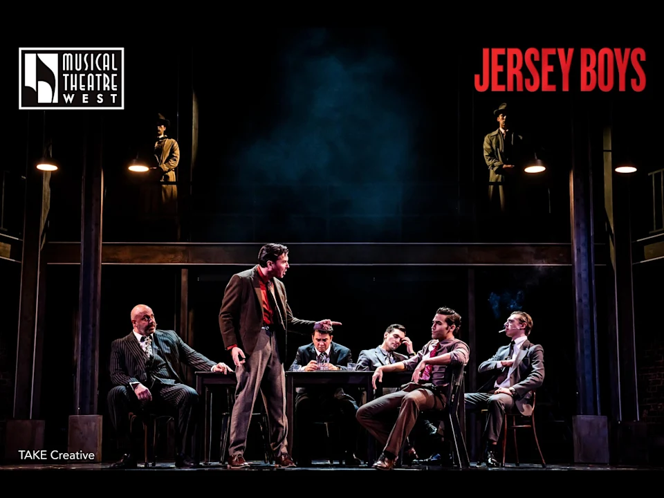 Stage production of "Jersey Boys" with six actors in suits, one standing and gesturing at a table while others sit in dim lighting.