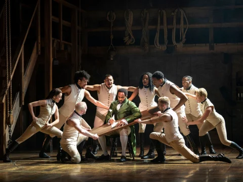 A group of performers in white outfits surrounds a man in a green coat sitting at a table, on stage with wooden backdrop and ropes, during a theatrical performance.