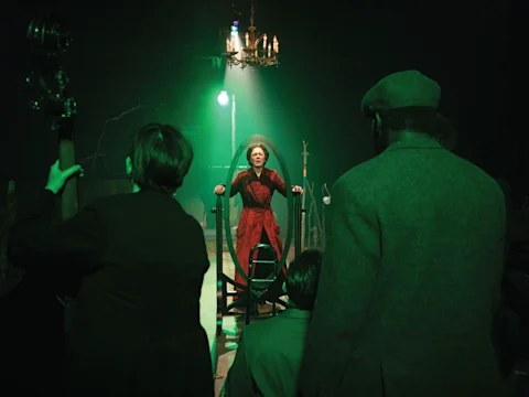 A woman in a red dress sits inside a large circular frame on stage, illuminated by green light, while several people watch her from the foreground.