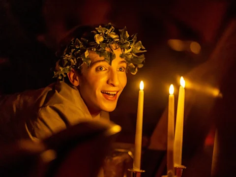 A person wearing a leafy wreath on their head smiles while looking at three lit candles in a dimly lit setting.