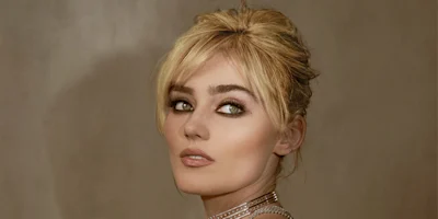 A woman with blonde hair styled up, wearing heavy eye makeup and a choker necklace, looks over her shoulder against a neutral background.