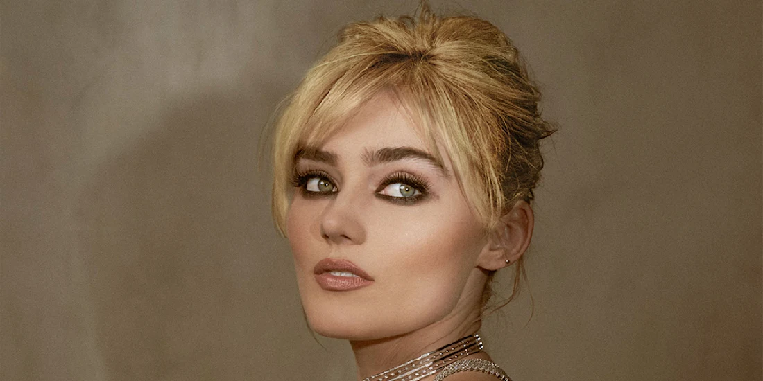 A woman with blonde hair styled up, wearing heavy eye makeup and a choker necklace, looks over her shoulder against a neutral background.