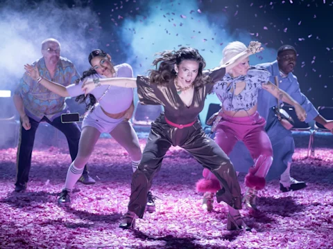 Five dancers perform energetically on a confetti-covered stage under dramatic lighting, with pink petals falling around them.