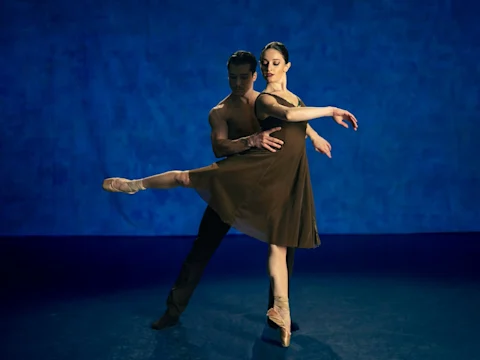 Two ballet dancers perform on stage; the male dancer supports the female dancer as she extends her leg, both poised against a blue background.