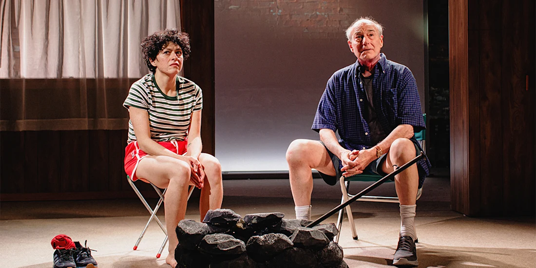 Two people sit on folding chairs indoors beside a pile of rocks; the woman wears a striped shirt and red shorts, the man wears a blue shirt and holds a cane.