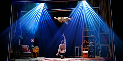 A person floats midair under blue stage lights as another person kneels below, reaching up in a room filled with furniture and props onstage.