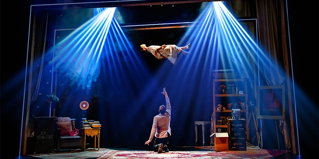 A person floats midair under blue stage lights as another person kneels below, reaching up in a room filled with furniture and props onstage.