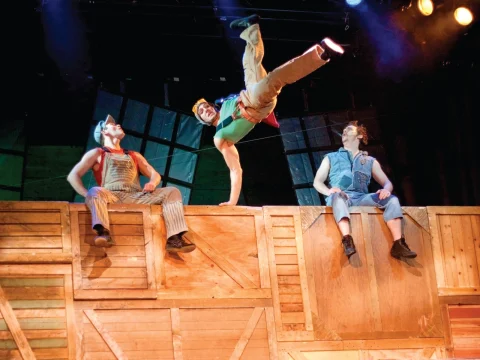 Three performers on stage, with one in mid-air doing a dynamic pose. They are surrounded by wooden crates under colorful stage lighting.