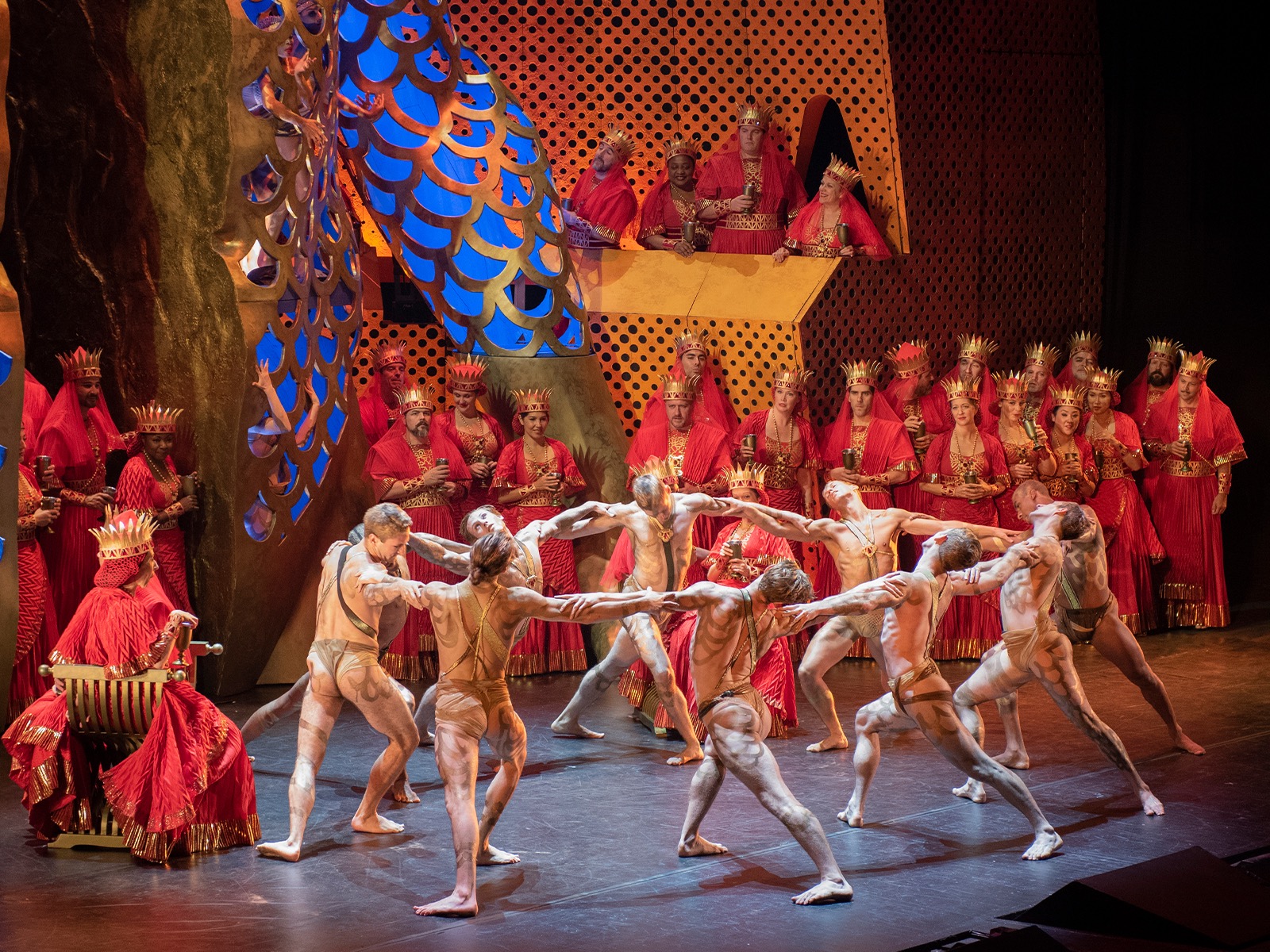 A group of dancers in nude-colored costumes perform on stage in front of seated and standing figures dressed in elaborate red robes and gold crowns.