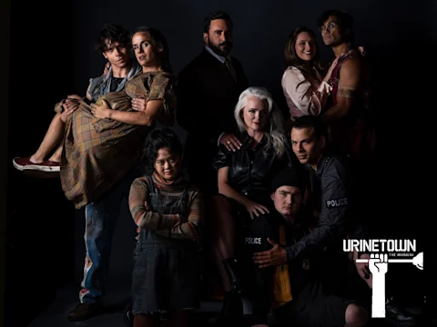 A group of nine people in theatrical costumes pose together against a black background with a "Urinetown The Musical" logo in the corner.