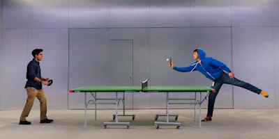Two people playing table tennis indoors; one serves while the other stands ready to receive.