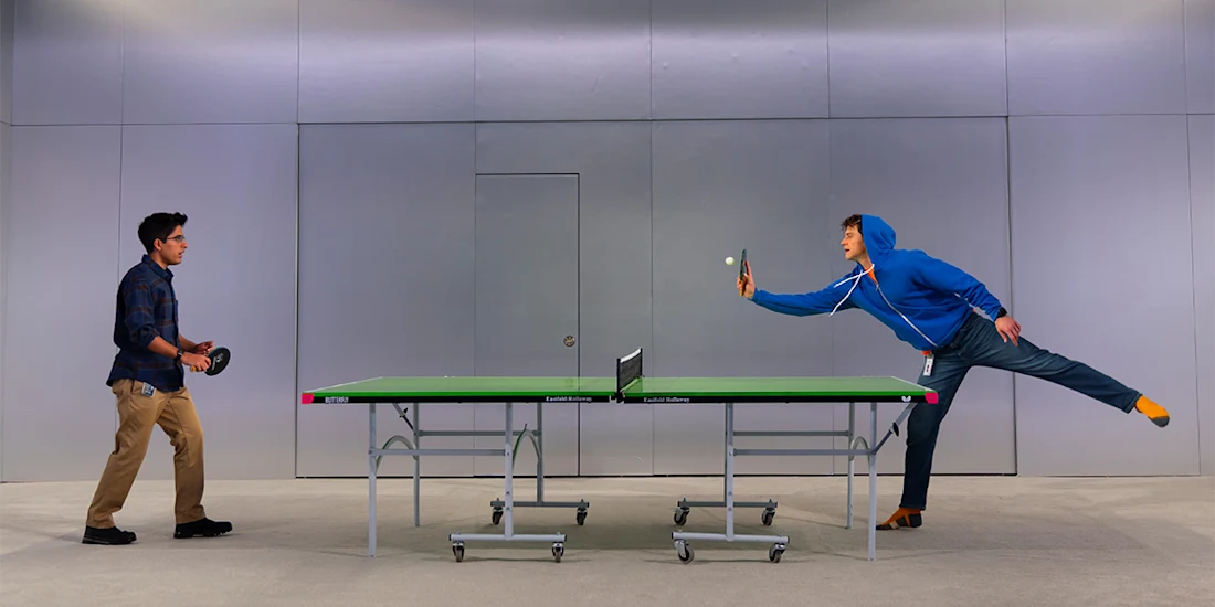 Two people playing table tennis indoors; one serves while the other stands ready to receive.