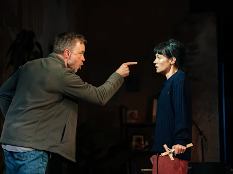 A man angrily points his finger at a woman holding a wooden cross, while she looks at him with a serious expression in a dimly lit room.