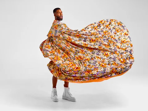A person in white boots poses with a large, colorful, orange floral fabric swirling around them against a plain white background.
