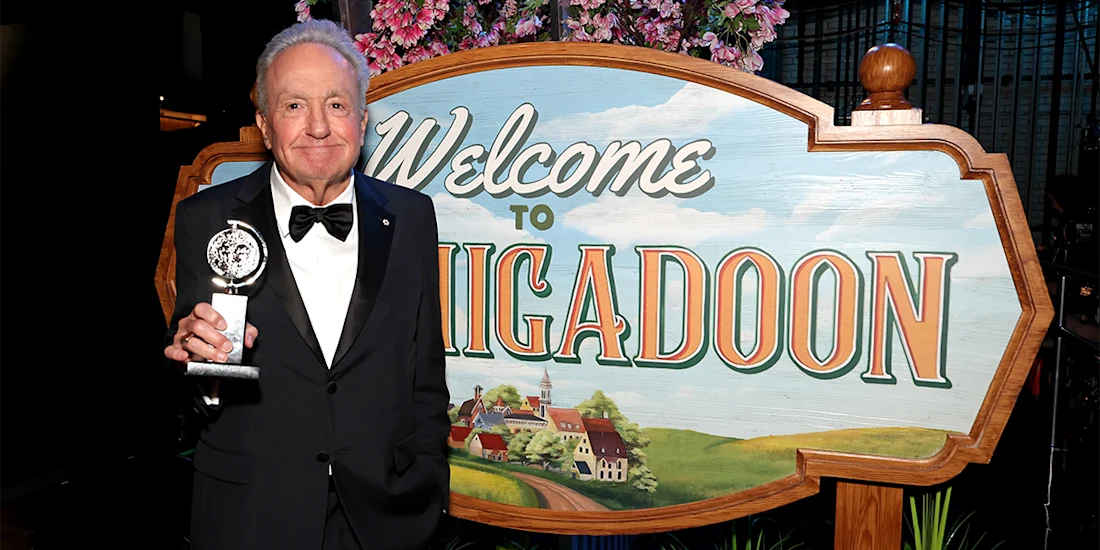 An older man in a tuxedo holds a trophy while standing beside a large "Welcome to Schmigadoon" sign decorated with flowers.
