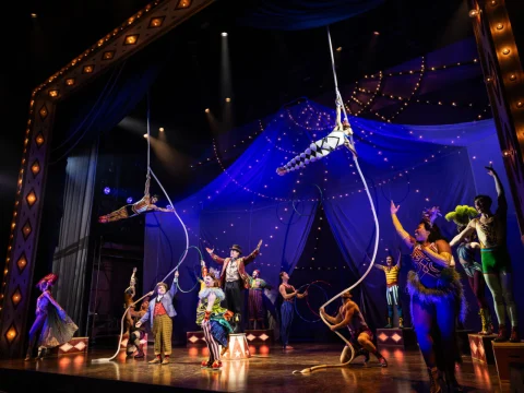 A vibrant circus performance with acrobats on ropes, a lively ensemble, and a backdrop of blue drapery and string lights on stage.