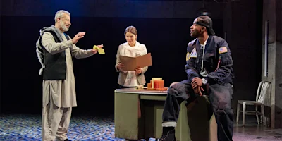 Three actors perform a scene on stage; one man gestures while holding paper, a woman stands behind a desk with a folder, and another man in a cap sits on the desk.