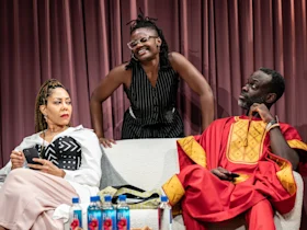 Three people on a couch; one standing and leaning over, smiling, while the seated individuals look in opposite directions. Bottles of water are on a table in front of them.