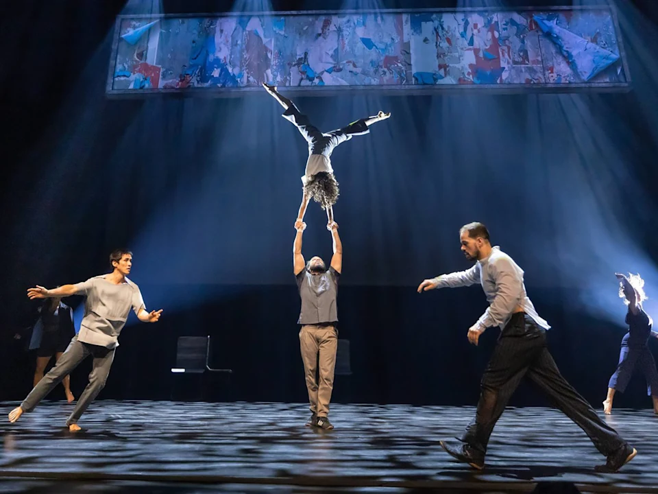 Five performers on stage; one balances upside down on another’s hands while others pose and move across a dimly lit set with a colorful mural overhead.