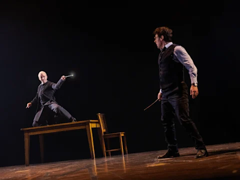 Two people face each other with wands drawn; one stands on a table aiming a glowing wand, while the other stands on the floor in a defensive stance.