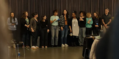 A group of eleven people stand in a line indoors near a curtain, some holding drinks or phones, engaging with each other and looking in different directions.
