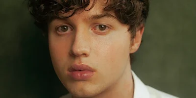 A person with curly brown hair and full lips looks directly at the camera against a softly blurred green background.