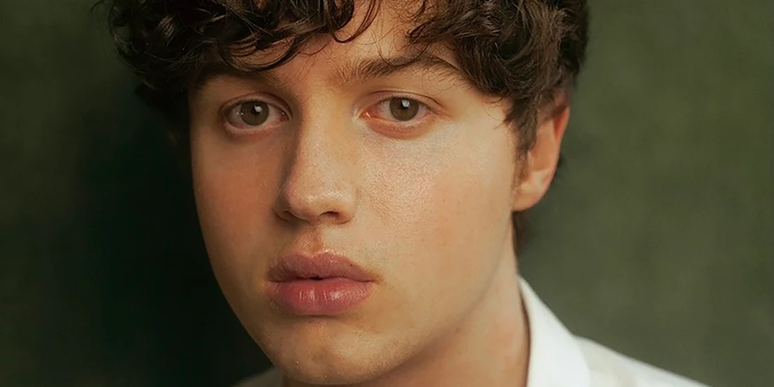 A person with curly brown hair and full lips looks directly at the camera against a softly blurred green background.