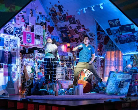 Two people energetically perform on a cluttered, colorful stage set resembling a decorated attic bedroom with posters, drawings, and photos covering the walls and ceiling.