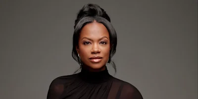 A woman with dark hair styled in an updo, wearing a black high-neck top, looks directly at the camera against a plain grey background.