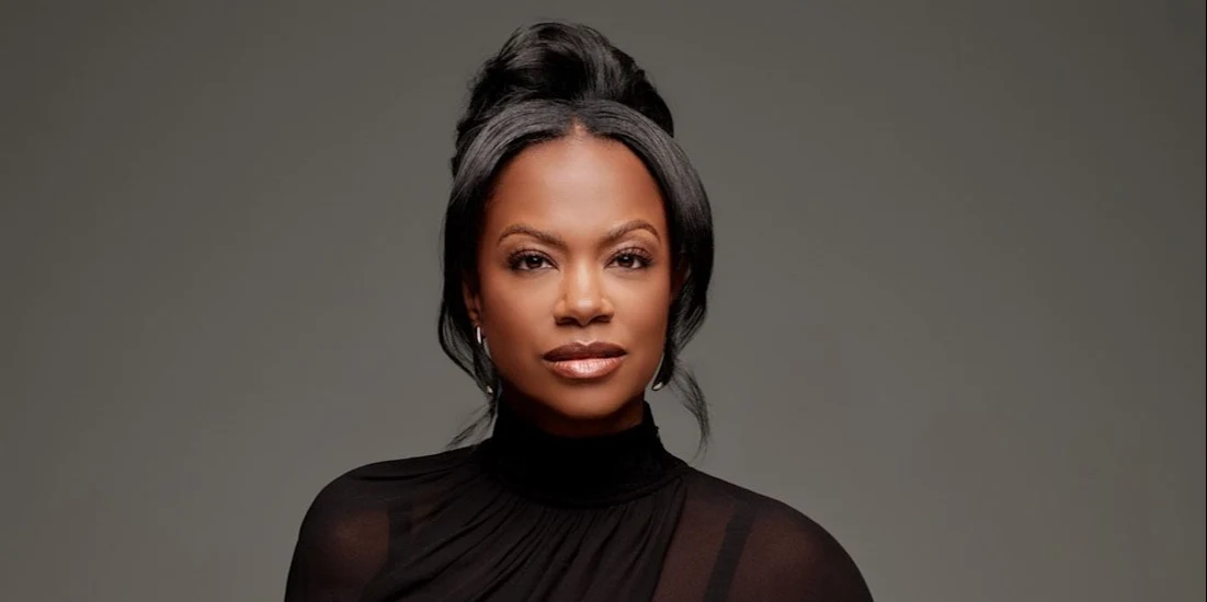 A woman with dark hair styled in an updo, wearing a black high-neck top, looks directly at the camera against a plain grey background.