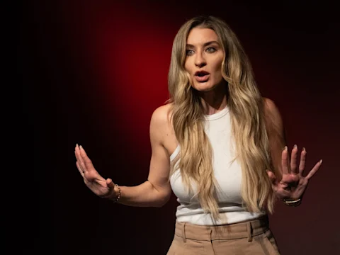 A woman with long blonde hair stands against a dark red background, wearing a white sleeveless top and tan pants, gesturing with both hands raised.