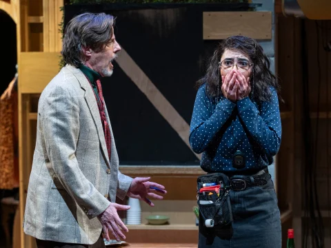 Two actors on stage. A man in a suit is speaking emphatically, and a woman in a polka dot shirt looks surprised, covering her mouth with her hands.