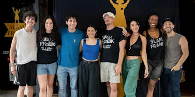 Eight people stand side by side, smiling at the camera in front of a “Slam Frank” banner. Some wear event t-shirts, and the group poses casually indoors.