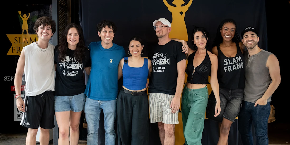 Eight people stand side by side, smiling at the camera in front of a “Slam Frank” banner. Some wear event t-shirts, and the group poses casually indoors.