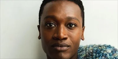 A person with short black hair looks directly at the camera, wearing a blue and white knit garment, against a plain white background.