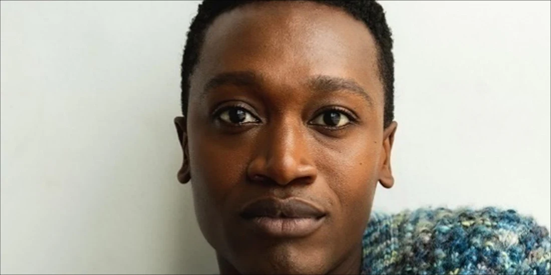 A person with short black hair looks directly at the camera, wearing a blue and white knit garment, against a plain white background.