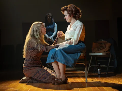 A woman kneels, holding the hand of another seated woman. A third person stands in the background. All are on a wooden floor in a room with muted lighting.