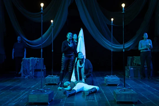 Actors perform a dramatic scene on stage with blue lighting, one person lying on the floor, others standing around, surrounded by draped fabric and tall lights.