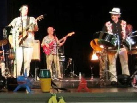 A band performing on stage with one person playing an electric guitar, another on steel drums, and others on drums and bass guitar. Toy figures are seen in the foreground.