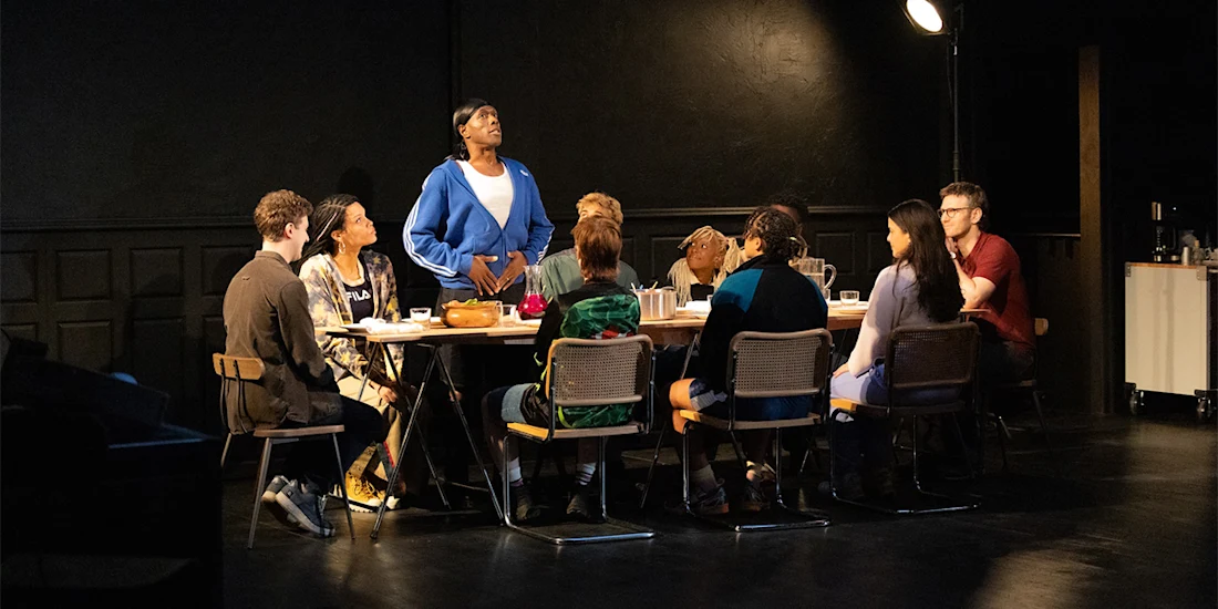 A diverse group of people sit around a long table, with one person standing and speaking while others listen attentively in a dimly lit room.