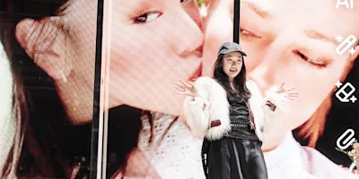 A woman in a white fur jacket and black hat poses with her hands up in front of a large screen displaying a close-up image of a face kissing a cheek.