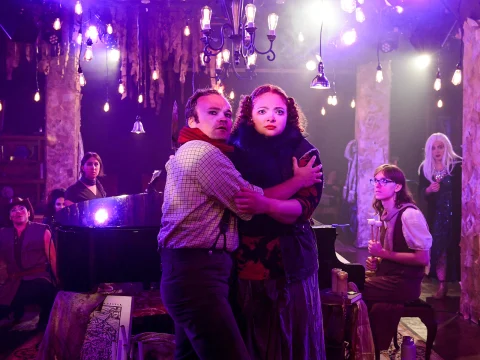 A man and a woman stand close together on stage surrounded by a piano and performers, under hanging lights with a dramatic atmosphere.