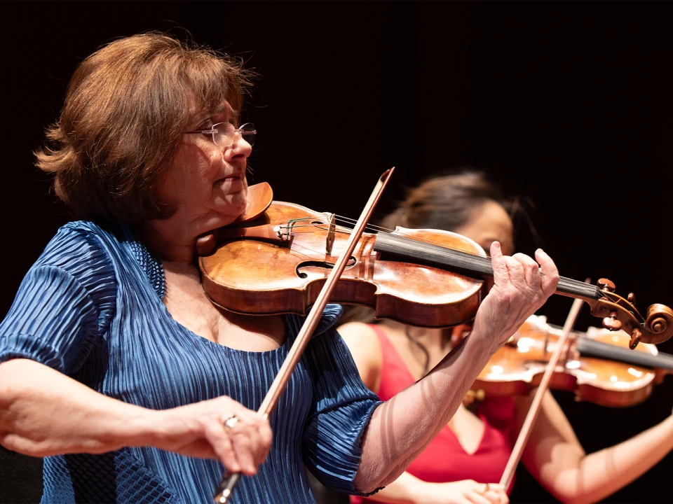 A woman wearing a blue top plays the violin passionately while another violinist is visible in the background.