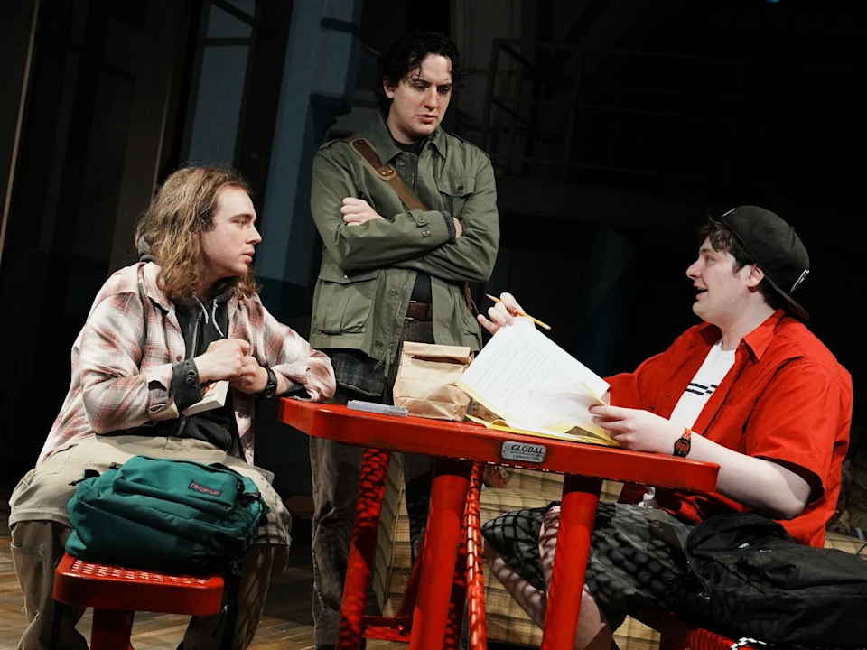 Production photo of Initiative in New York, showing three people sit and stand around a red table, engaged in discussion; one is holding a pencil and paper, another has a backpack, and the third stands with folded arms.