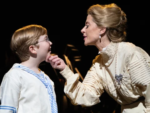 A woman in a white Victorian-style dress touches the chin of a young boy wearing glasses and a sailor outfit on stage.