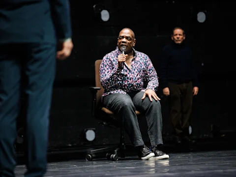 A man in a patterned shirt sits on an office chair holding a microphone, with two other people standing and sitting in the background on a stage.
