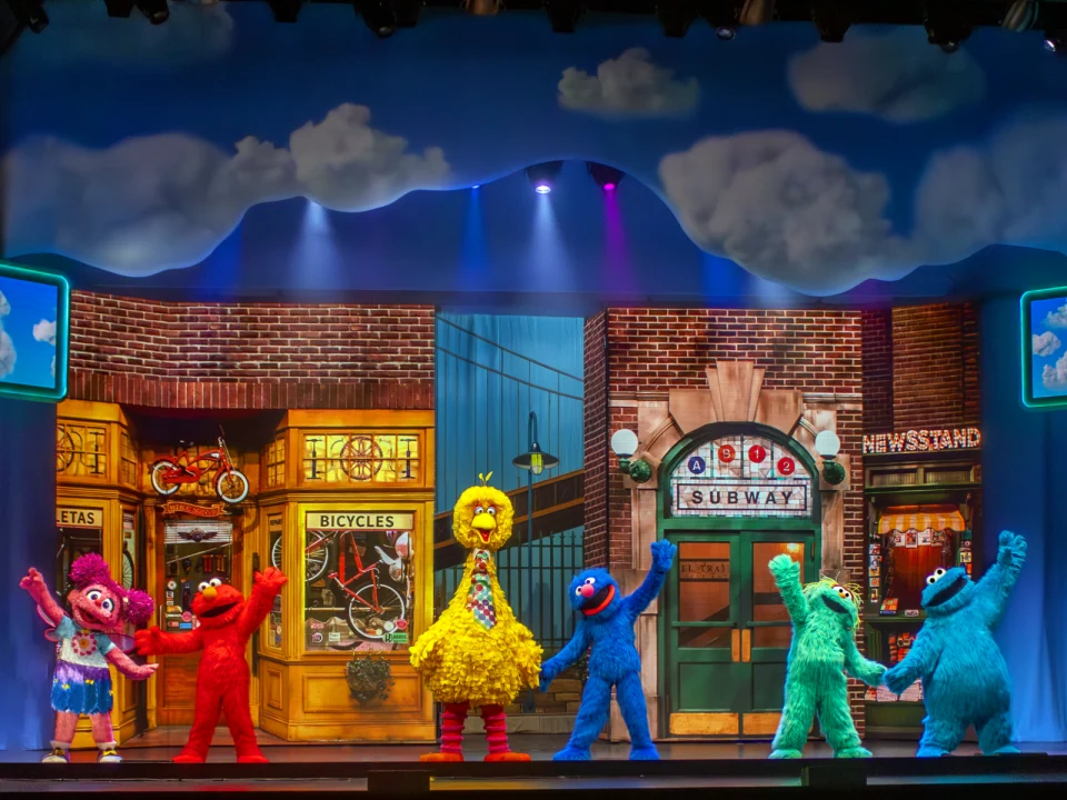 Costumed characters are performing on stage with a colorful backdrop featuring a shop, a subway entrance, and floating clouds.